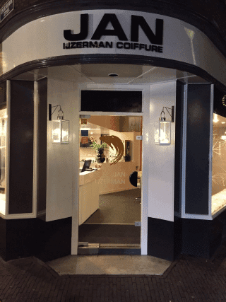 De verlichte gevel van Jan IJzerman Coiffure met de openstaande deuren naar de salon