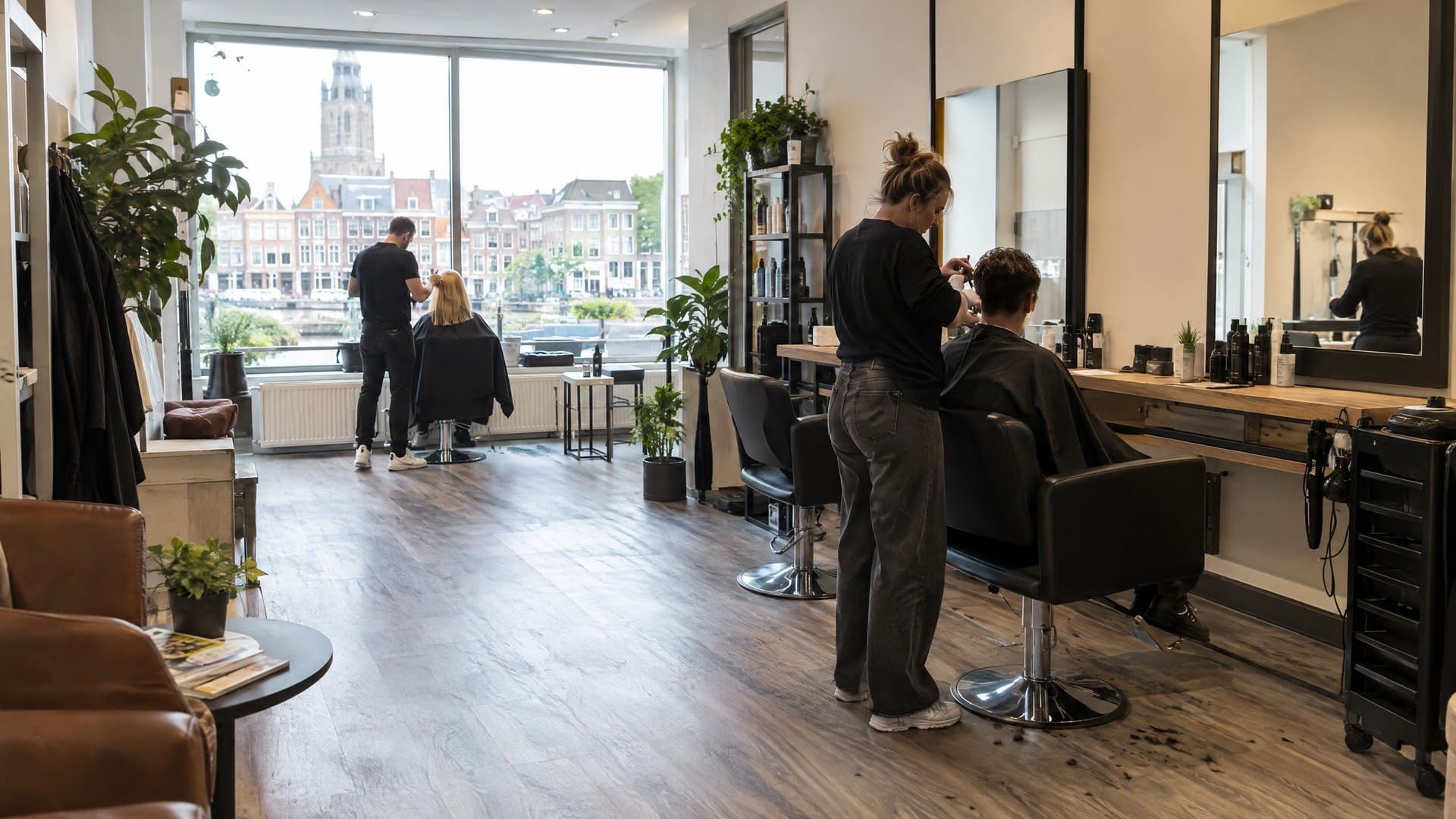 Een lichte kapsalon met houten vloer, zwarte stoelen en grote ramen met uitzicht op een historische Nederlandse stadskade